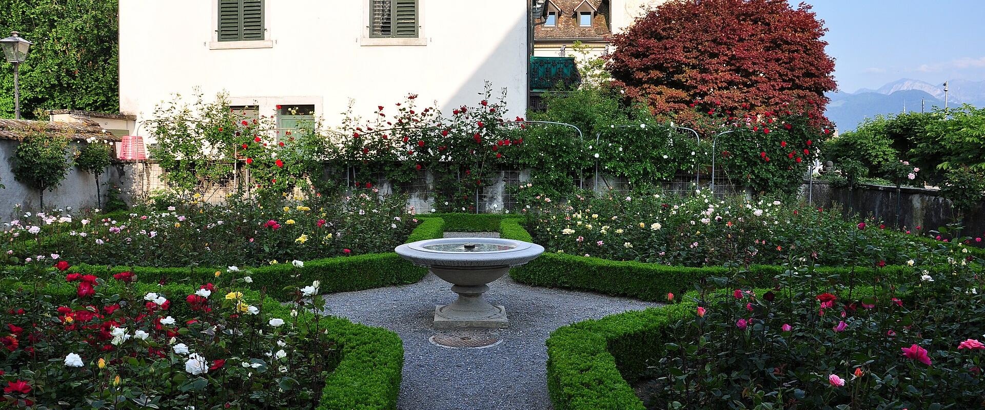 A formal rose garden with a central stone fountain, gravel paths, and manicured hedges, set against a white building with green shutters and a view of distant mountains.