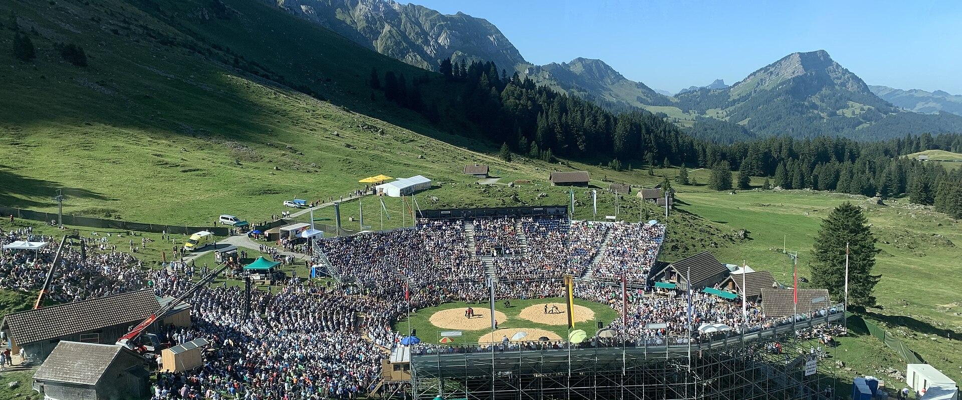 An aerial view of a large outdoor Schwinget (Swiss wrestling) arena filled with spectators, set on a grassy mountain plateau with steep rocky peaks in the background.