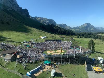 An aerial view of a large outdoor Schwinget (Swiss wrestling) arena filled with spectators, set on a grassy mountain plateau with steep rocky peaks in the background.