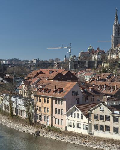 A panoramic view of a historic European city center situated on a peninsula formed by a river, featuring a prominent cathedral spire, a stone arch bridge, and a modern truss bridge in the distance, viewed from an elevated park with trees in the foreg