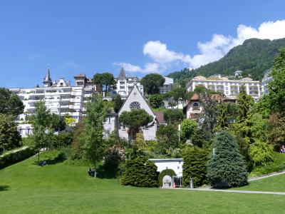 A grassy park slope with manicured trees and bushes in the foreground, leading up to a small stone chapel-like building, with large white hotels and a forested hillside in the background under a blue sky.