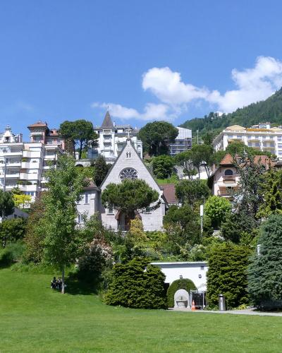 A grassy park slope with manicured trees and bushes in the foreground, leading up to a small stone chapel-like building, with large white hotels and a forested hillside in the background under a blue sky.