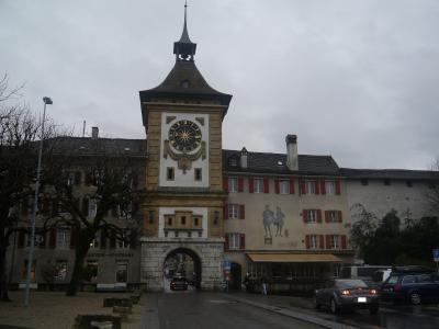 A stone clock tower with an arched gateway (the Berntor) in a European town, featuring a pharmacy sign reading "BERNTOR-APOTHEKE" and a mural of two figures on the adjacent building.