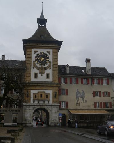 A stone clock tower with an arched gateway (the Berntor) in a European town, featuring a pharmacy sign reading "BERNTOR-APOTHEKE" and a mural of two figures on the adjacent building.