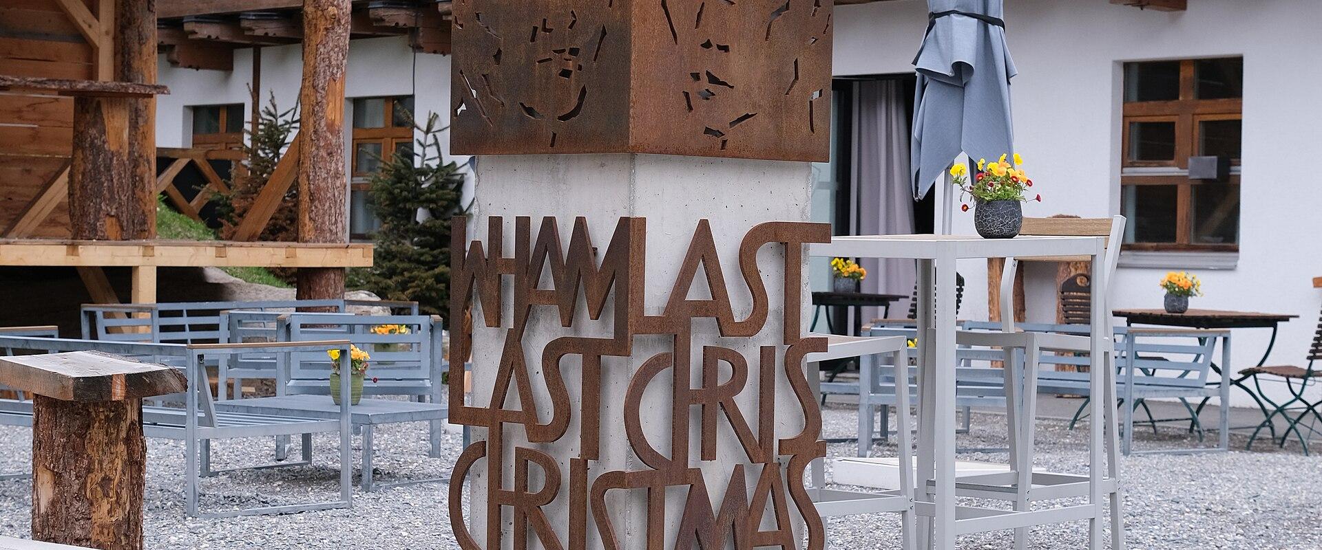 A concrete and corten steel monument featuring the text "WHAM! LAST CHRISTMAS" and "Saas-Fee 1984" standing in front of a wooden chalet-style building with outdoor seating.