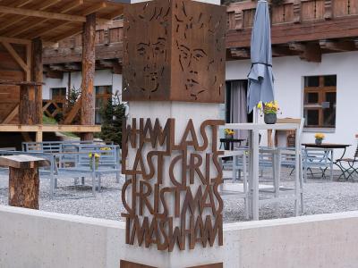 A concrete and corten steel monument featuring the text "WHAM! LAST CHRISTMAS" and "Saas-Fee 1984" standing in front of a wooden chalet-style building with outdoor seating.