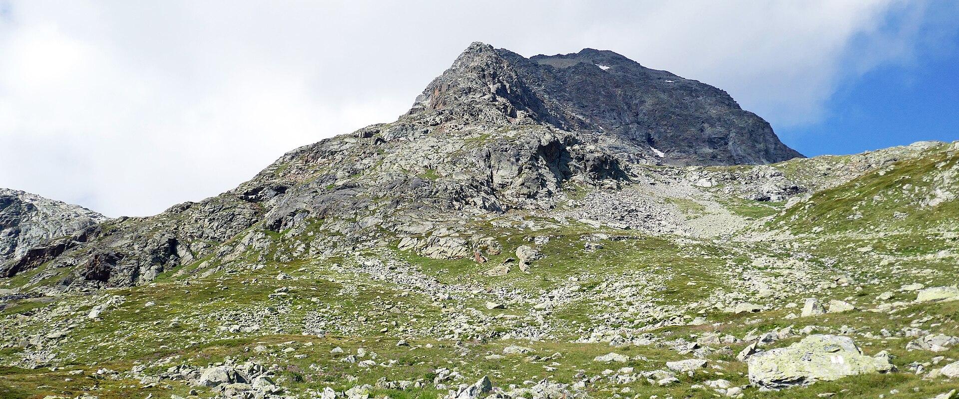 A rocky mountain peak rising above a grassy, scree-covered alpine slope under a cloudy sky.