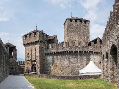 A view of the interior courtyard of a medieval stone fortress featuring a tall square tower, a smaller tower with a wooden balcony, and a white event tent set up on the grass.