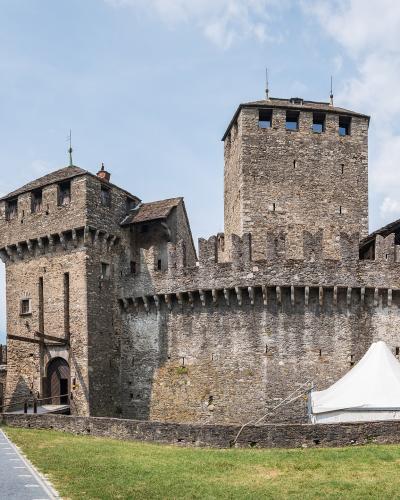 A view of the interior courtyard of a medieval stone fortress featuring a tall square tower, a smaller tower with a wooden balcony, and a white event tent set up on the grass.