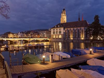 A twilight view of the Limmat River in Zürich featuring the Wasserkirche (Water Church) on the right bank, with the Fraumünster church tower visible behind it, the Münsterbrücke bridge spanning the river, and covered boats moored at a wooden dock