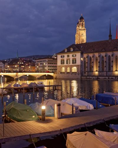 A twilight view of the Limmat River in Zürich featuring the Wasserkirche (Water Church) on the right bank, with the Fraumünster church tower visible behind it, the Münsterbrücke bridge spanning the river, and covered boats moored at a wooden dock