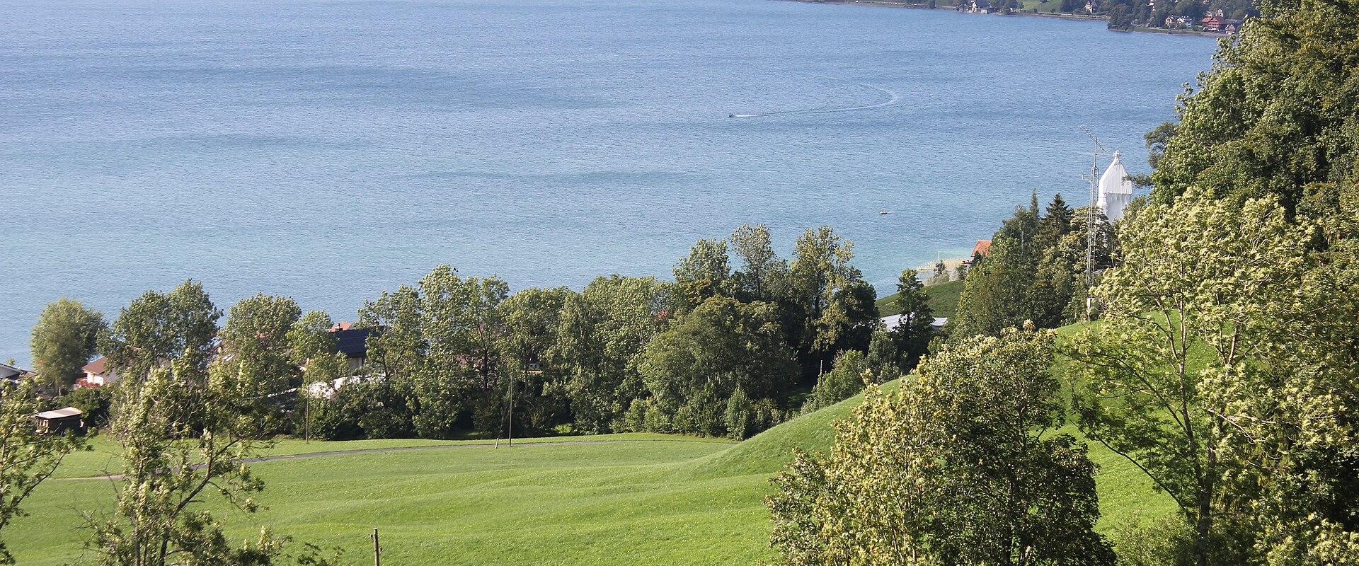 A high-angle landscape view of a large lake with a town along the far shore, green hills in the foreground, and a white chapel on a hill to the right.