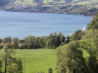 A high-angle landscape view of a large lake with a town along the far shore, green hills in the foreground, and a white chapel on a hill to the right.