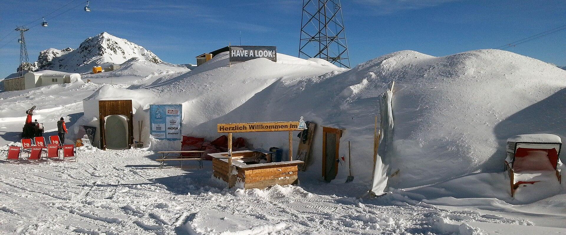 A snowy outdoor scene featuring a temporary ice or snow structure with wooden entrances, red lounge chairs, and signs reading "HAVE A LOOK!" and "Herzlich Willkommen Im" (German for "Welcome In"), with a ski lift and mountain in the background.