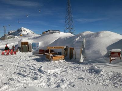 A snowy outdoor scene featuring a temporary ice or snow structure with wooden entrances, red lounge chairs, and signs reading "HAVE A LOOK!" and "Herzlich Willkommen Im" (German for "Welcome In"), with a ski lift and mountain in the background.