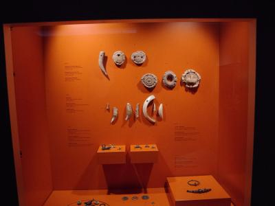 A museum display case with an orange background containing various ancient silver artifacts, including pendants, rings, and coins, with descriptive text panels on the left and right walls.