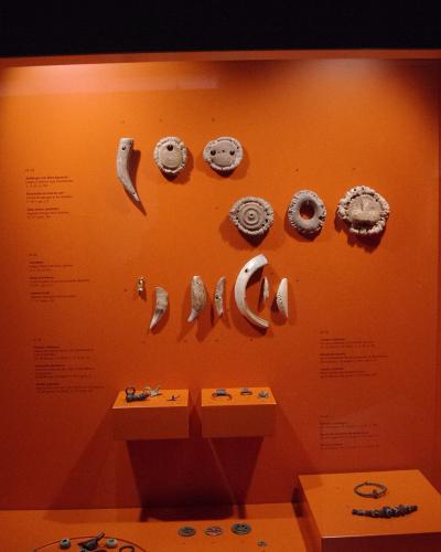 A museum display case with an orange background containing various ancient silver artifacts, including pendants, rings, and coins, with descriptive text panels on the left and right walls.