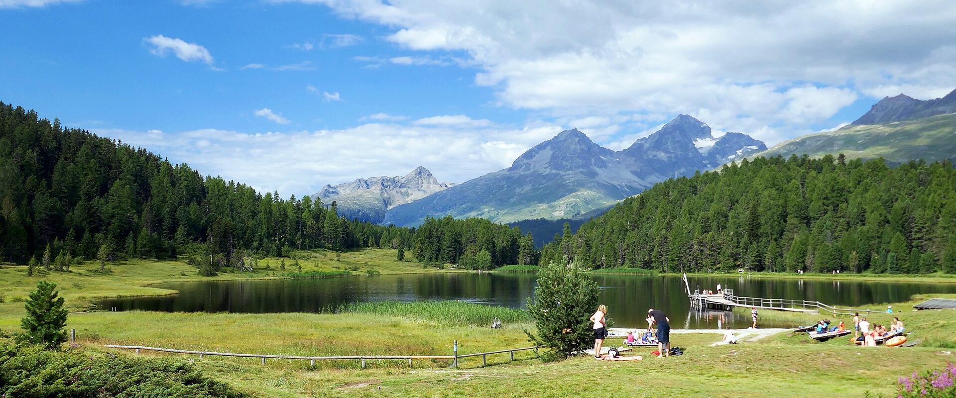 A panoramic view of a calm alpine lake surrounded by dense coniferous forests and distant rocky mountain peaks, with people relaxing on the grassy shore and a wooden pier extending into the water.