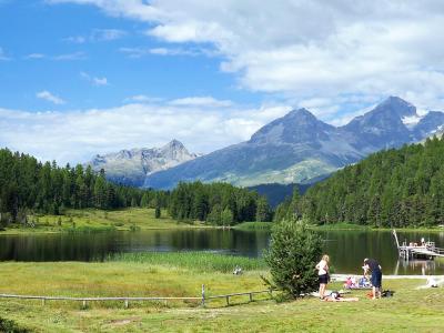 A panoramic view of a calm alpine lake surrounded by dense coniferous forests and distant rocky mountain peaks, with people relaxing on the grassy shore and a wooden pier extending into the water.