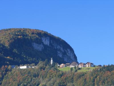 A distant view of a village nestled on a green hillside at the base of a large, steep mountain with a prominent rock face, featuring a church with a white tower and several buildings.
