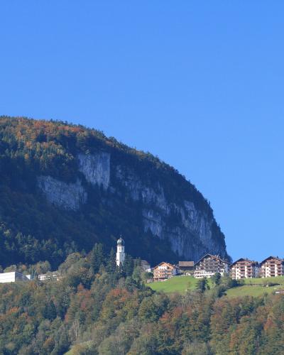 A distant view of a village nestled on a green hillside at the base of a large, steep mountain with a prominent rock face, featuring a church with a white tower and several buildings.