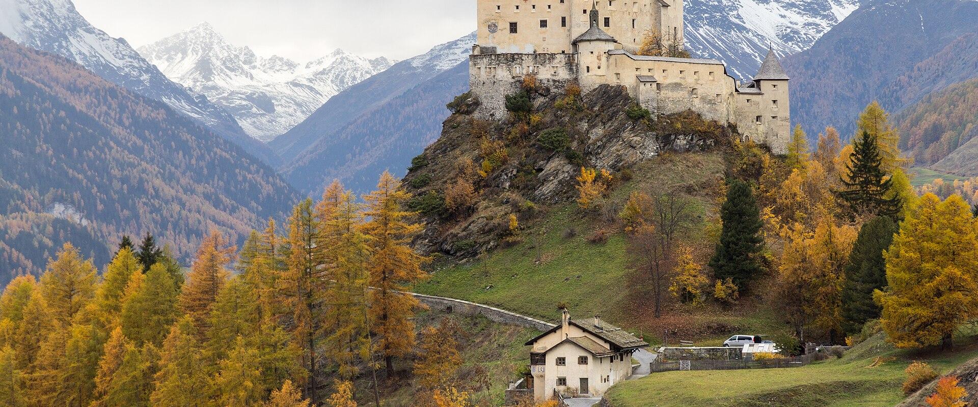 Tarasp Castle perched on a rocky hill, surrounded by autumn-colored trees and snow-capped mountains in the background.