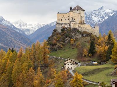 Tarasp Castle perched on a rocky hill, surrounded by autumn-colored trees and snow-capped mountains in the background.