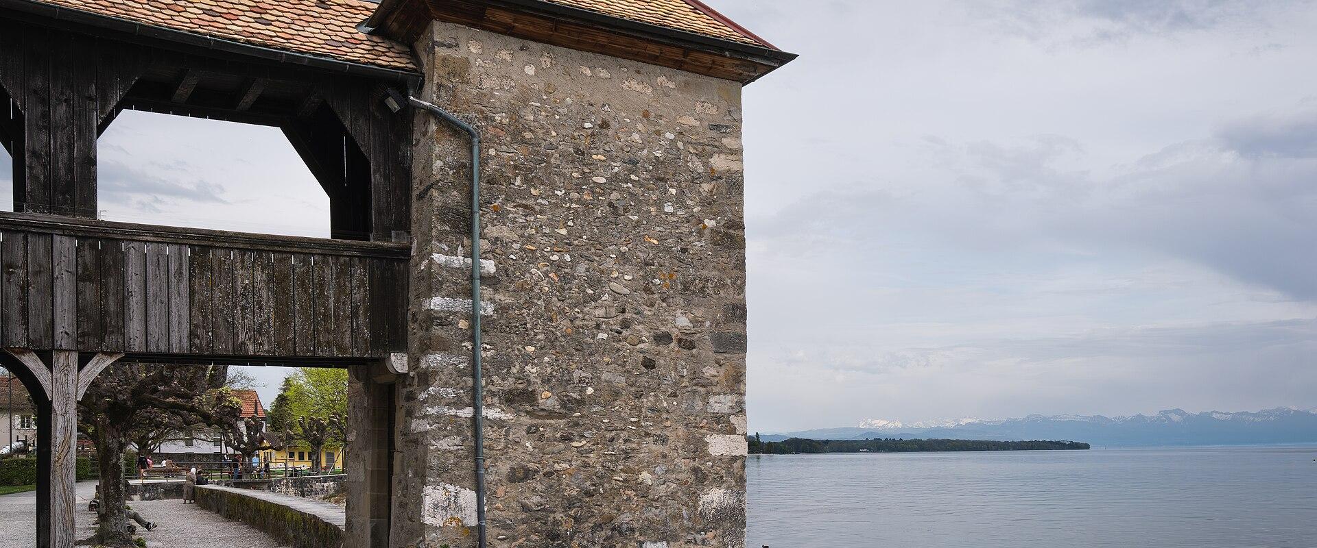A stone tower with a tiled roof and a wooden covered walkway attached to it, situated on the shore of a large lake with mountains in the distance.