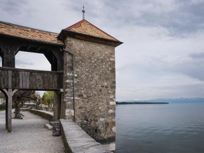 A stone tower with a tiled roof and a wooden covered walkway attached to it, situated on the shore of a large lake with mountains in the distance.