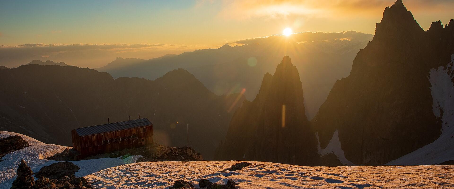 A wooden mountain hut situated on a snowy ridge at sunrise, with dramatic jagged peaks and layered mountain ranges in the background.