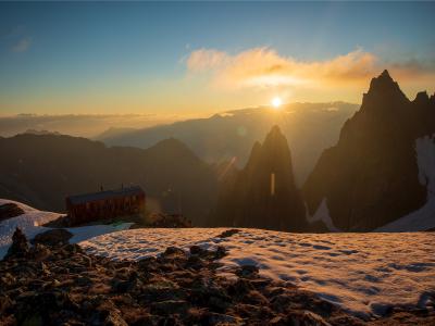 A wooden mountain hut situated on a snowy ridge at sunrise, with dramatic jagged peaks and layered mountain ranges in the background.