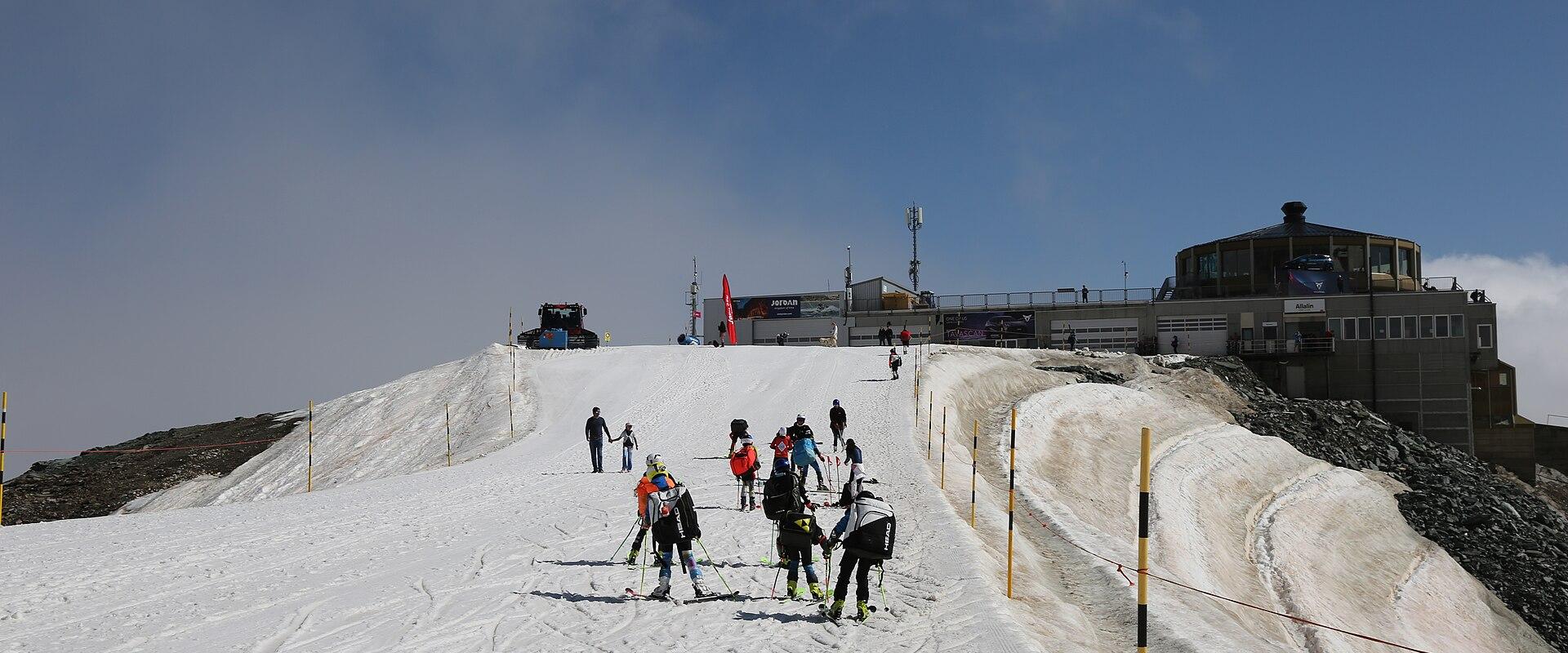 A group of skiers on a snowy slope leading up to a mountain station building with a rotating restaurant structure on the right.