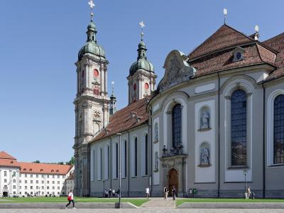 A large Baroque-style cathedral with twin green-domed towers and a long nave, situated next to a white administrative building with a red roof under a clear blue sky.