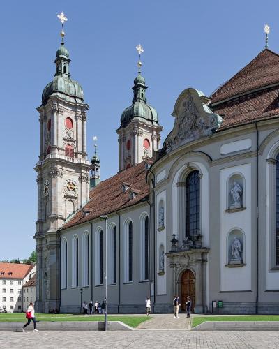 A large Baroque-style cathedral with twin green-domed towers and a long nave, situated next to a white administrative building with a red roof under a clear blue sky.