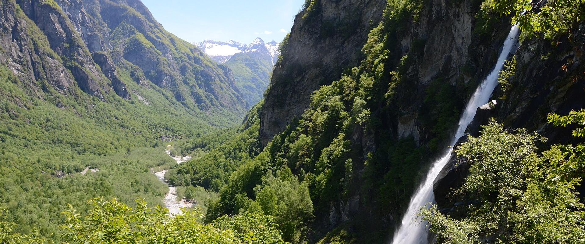 A tall, narrow waterfall cascading down a steep, rocky cliff face into a lush green valley with a river and distant snow-capped mountains.