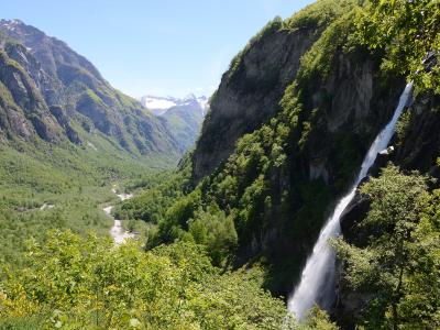A tall, narrow waterfall cascading down a steep, rocky cliff face into a lush green valley with a river and distant snow-capped mountains.