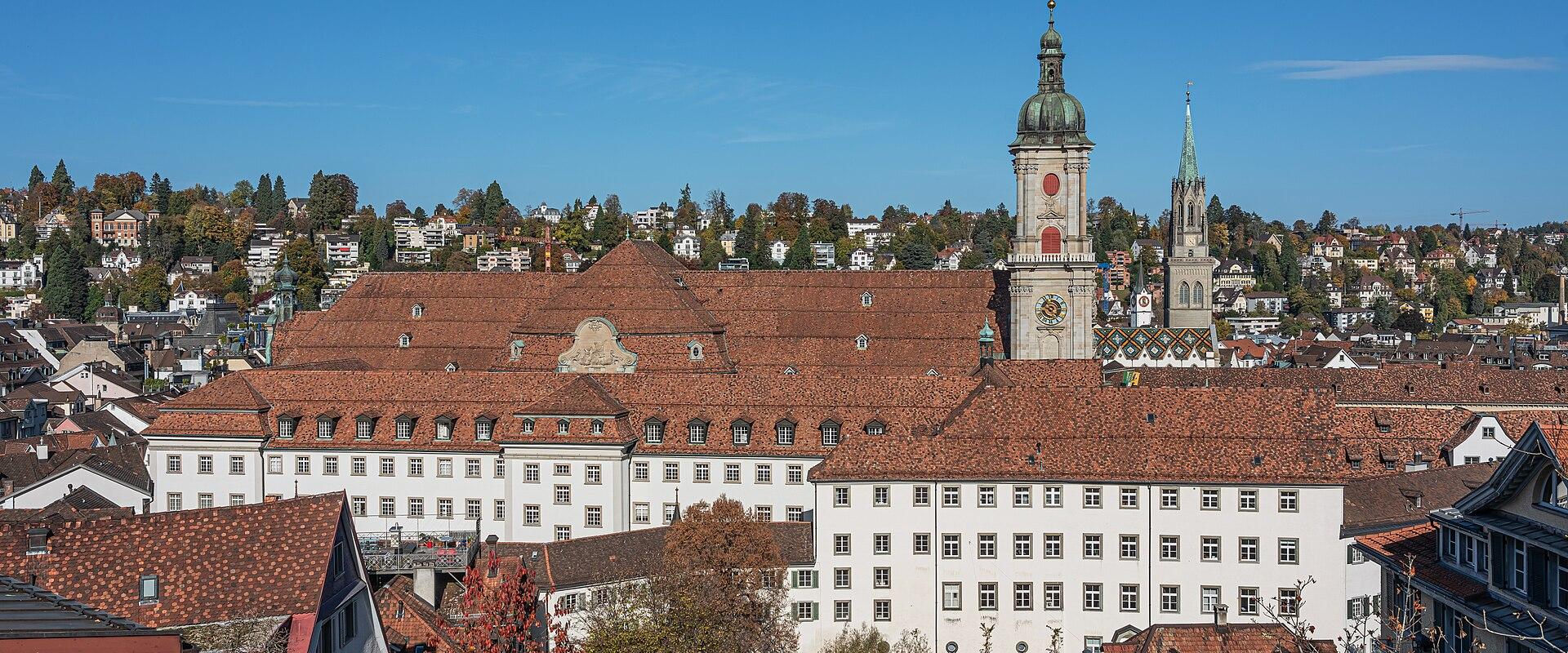 An exterior view of the Abbey of St. Gallen complex, showing the large white buildings with red-tiled roofs, the distinctive green-domed abbey tower, and the adjacent cathedral spire against a blue sky.