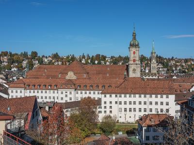 An exterior view of the Abbey of St. Gallen complex, showing the large white buildings with red-tiled roofs, the distinctive green-domed abbey tower, and the adjacent cathedral spire against a blue sky.
