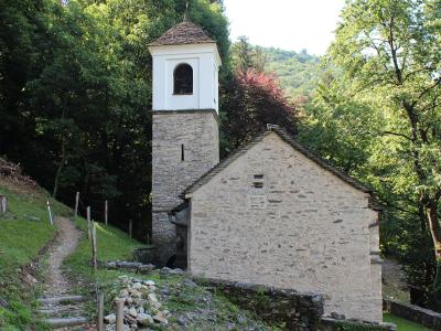 A stone church with a white bell tower and tiled roof, situated on a grassy hillside next to a dirt path with wooden posts and rope fencing, surrounded by dense green trees.