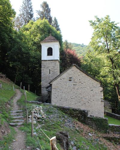 A stone church with a white bell tower and tiled roof, situated on a grassy hillside next to a dirt path with wooden posts and rope fencing, surrounded by dense green trees.