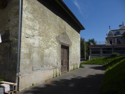 A weathered stone building with a wooden door and a paved path leading uphill toward a large villa-style house under a blue sky.