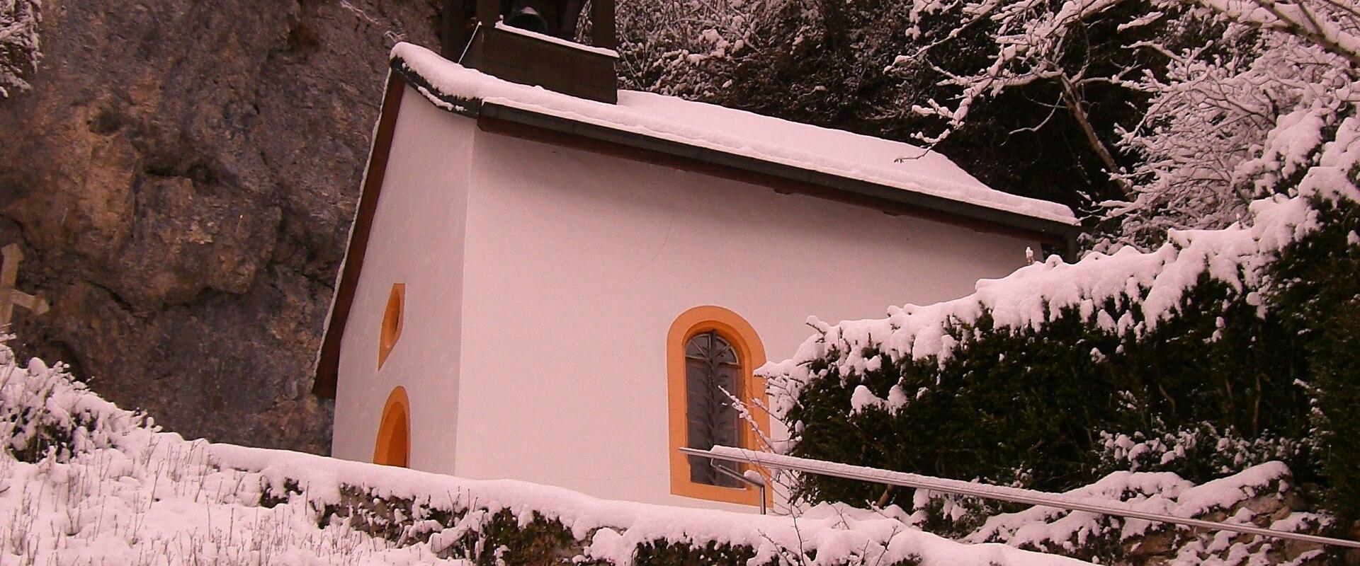 A small white chapel with a bell tower and orange-trimmed windows, situated next to a rocky cliff face and surrounded by snow-covered vegetation and a stone wall.