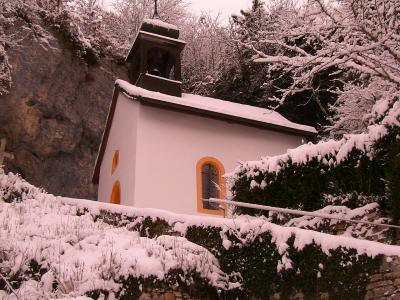 A small white chapel with a bell tower and orange-trimmed windows, situated next to a rocky cliff face and surrounded by snow-covered vegetation and a stone wall.