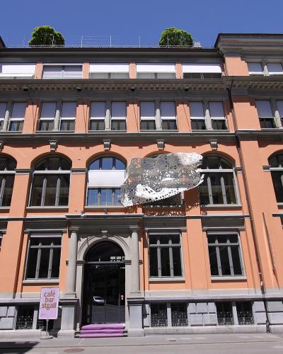 A peach-colored historic building facade with a large white lace installation hanging from the second-story windows and a sign for "café bar st.gall" near the entrance.