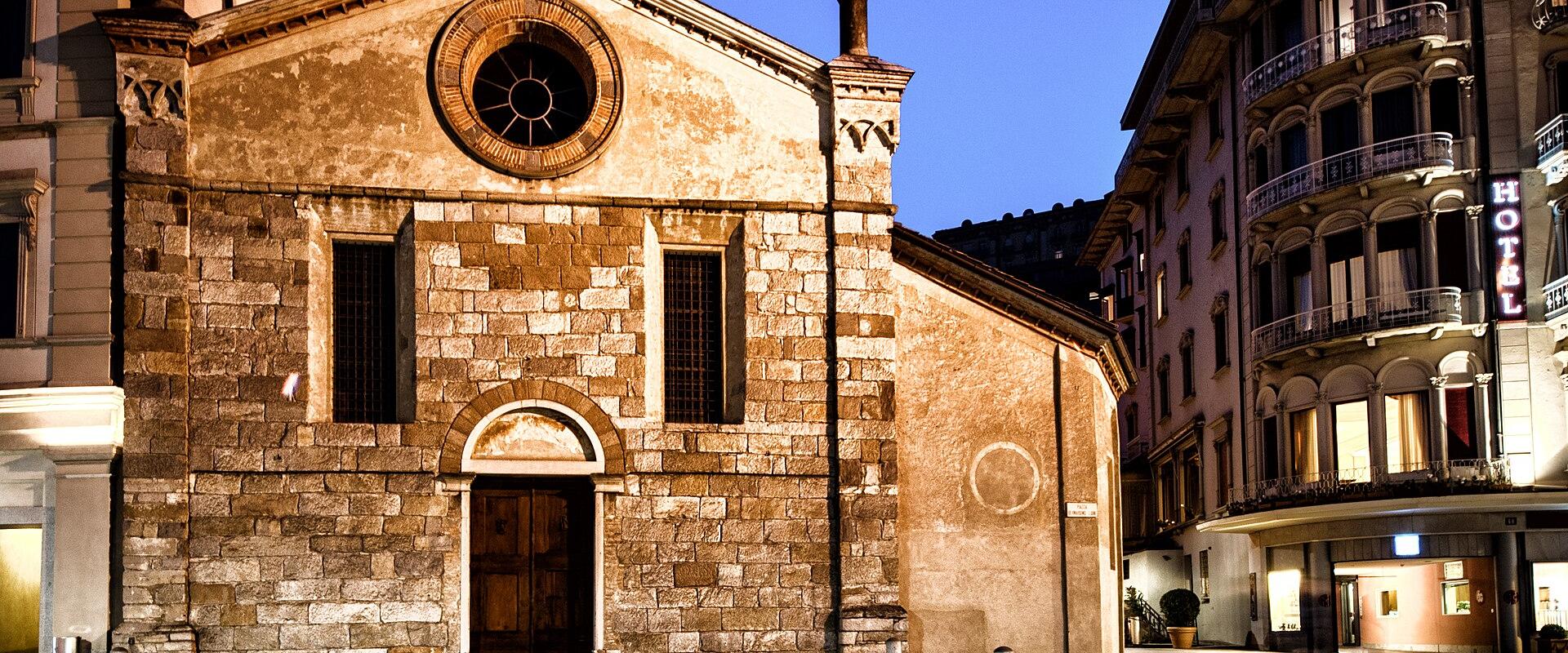 A stone church facade with a central rose window and arched doorway, situated in a paved square at twilight next to a building with a vertical "HOTEL" sign.