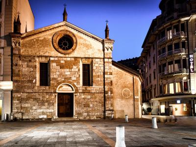 A stone church facade with a central rose window and arched doorway, situated in a paved square at twilight next to a building with a vertical "HOTEL" sign.