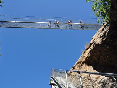 A low-angle view looking up at a metal mesh suspension footbridge spanning a rocky gorge, with four people walking across it against a clear blue sky.