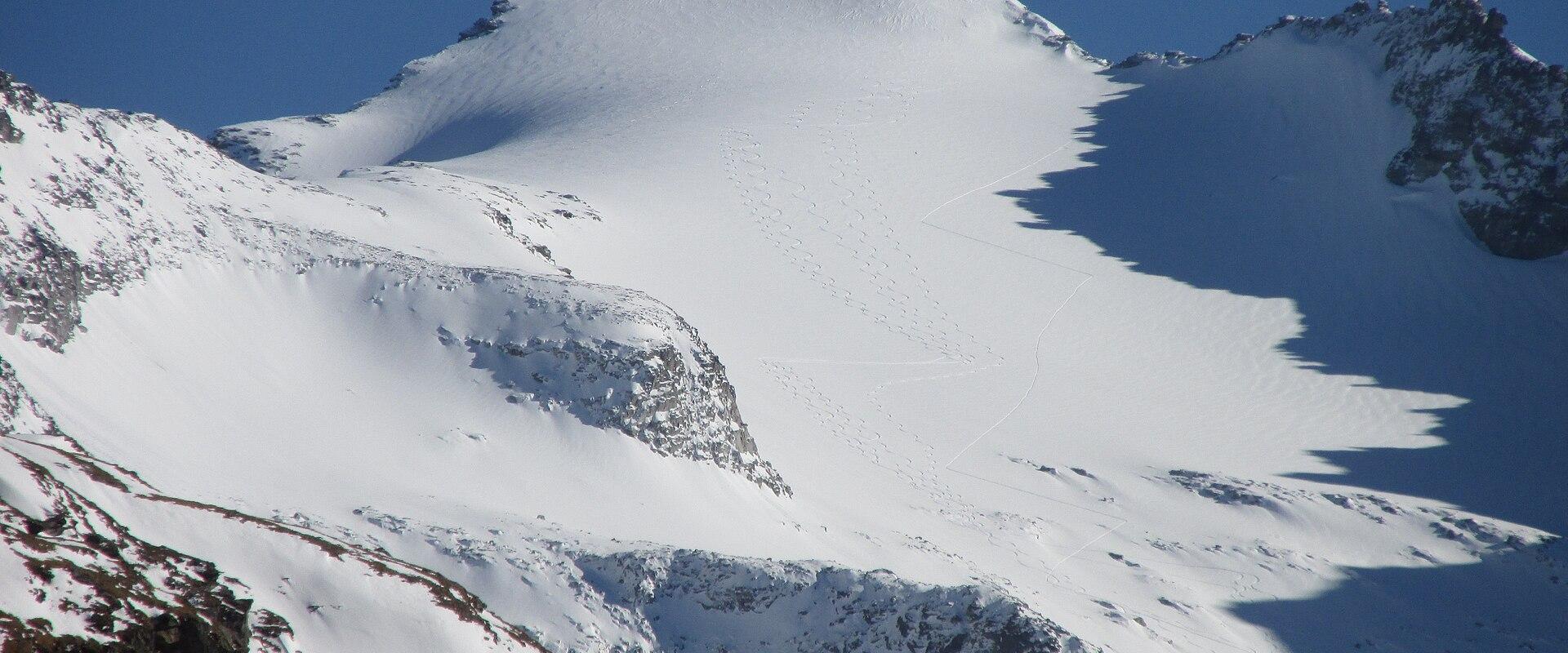 A snow-covered mountain peak with rocky outcrops under a clear blue sky, showing ski tracks on the slope.