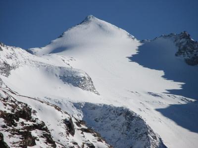 A snow-covered mountain peak with rocky outcrops under a clear blue sky, showing ski tracks on the slope.