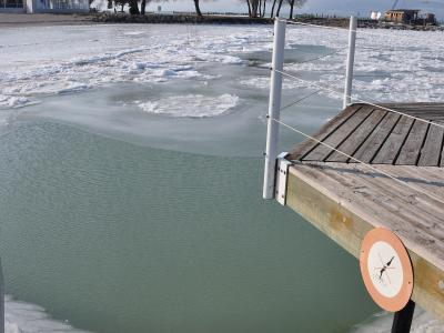 A frozen lake scene showing a wooden pier with white posts and a circular orange sign, with ice floes on the water and bare trees on the shore in the background.
